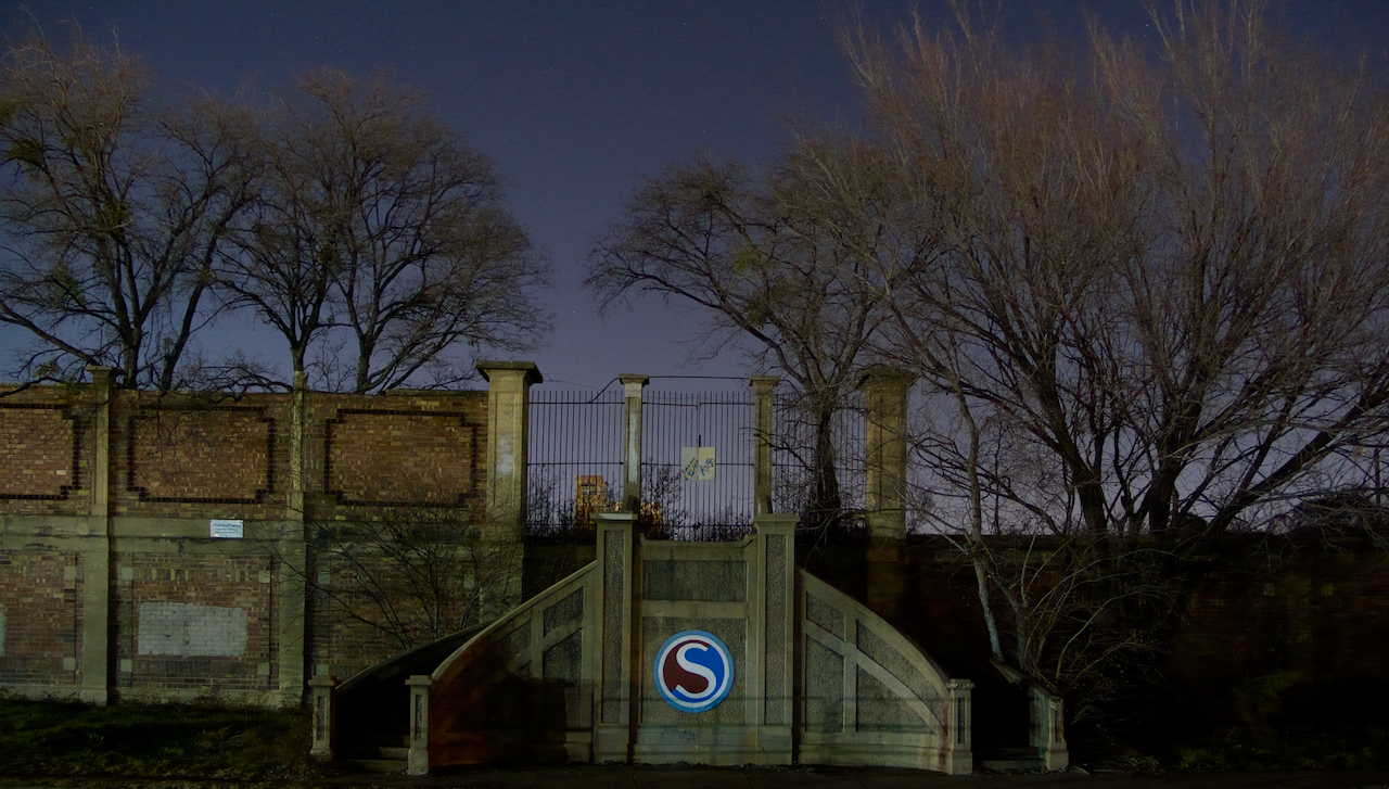 Dark outdoor scene with a staircase and wall featuring a logo, trees in the background. The abandoned Swift Meatpacking headquarters entrance.